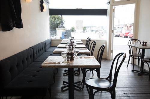 Interior of restaurant with black wall seating, square wooden tables and  wooden chairs on right. Large window at end of tables